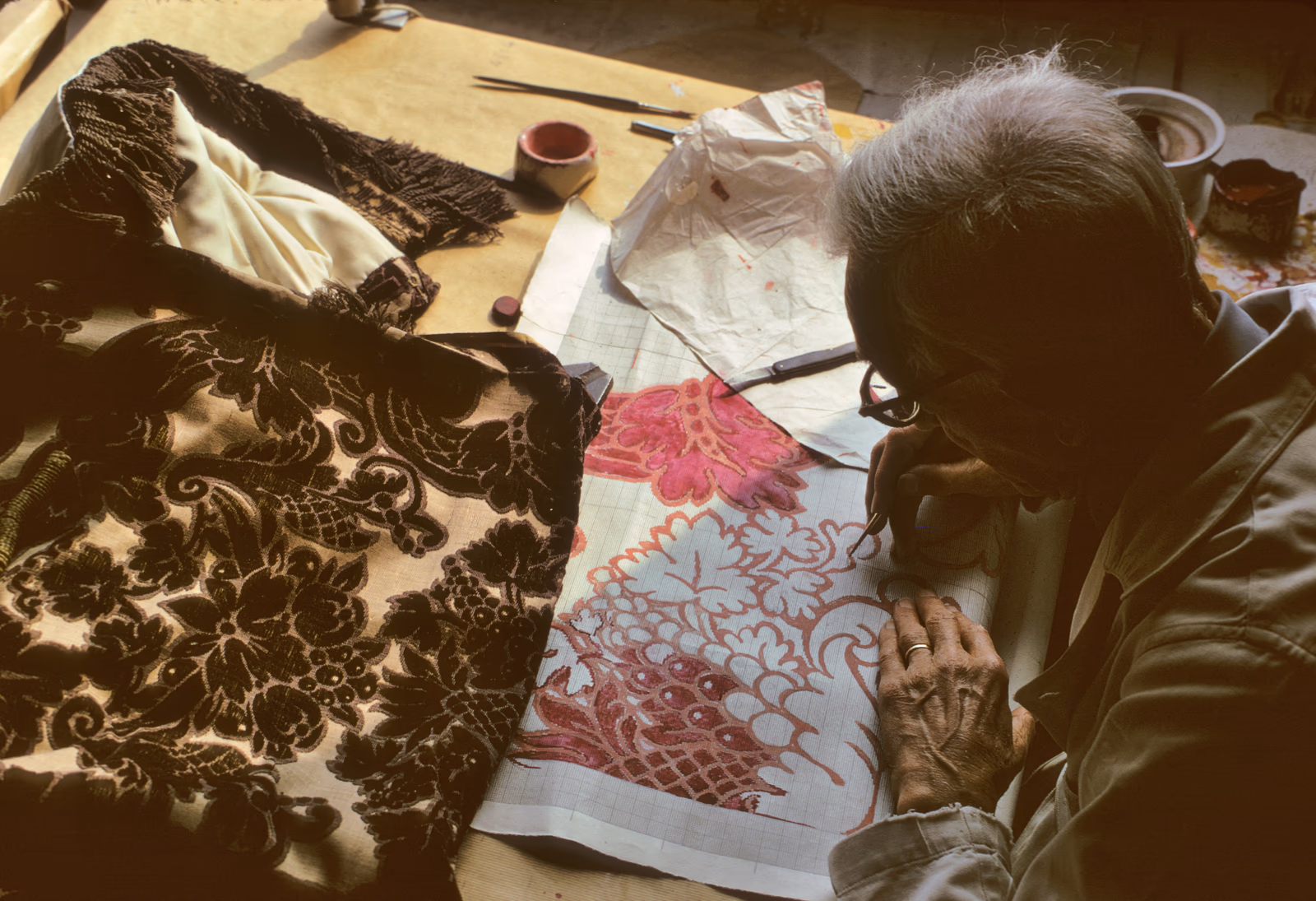 Artisan peignant un motif floral rouge sur tissu, entouré d'étoffes anciennes